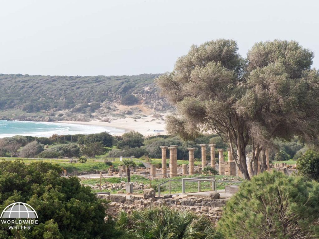 Looking over the remains of a Roman town. There are big trees to the right, and behind the town is the sea, surrounded by beach and cliffs.